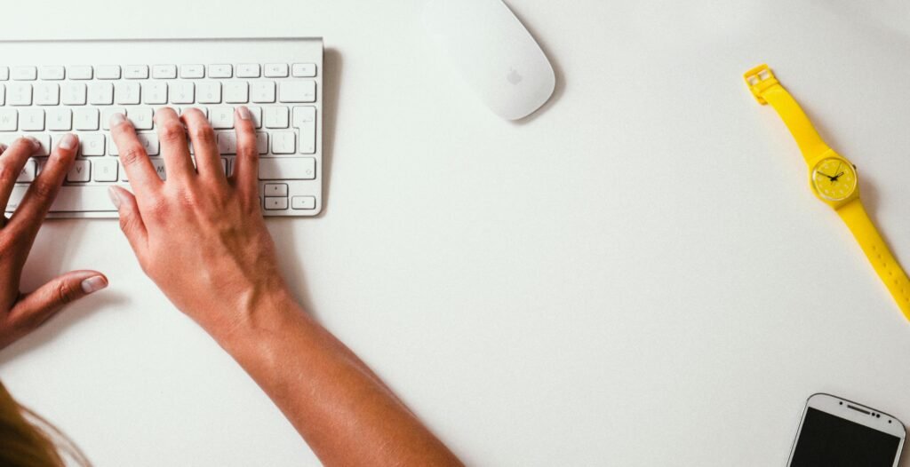 Close-up of hands typing on a keyboard with a yellow watch on the desk
