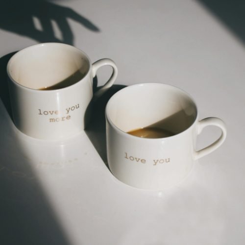 Pair of ceramic mugs with 'love you' text, casting artistic shadows on a white surface.